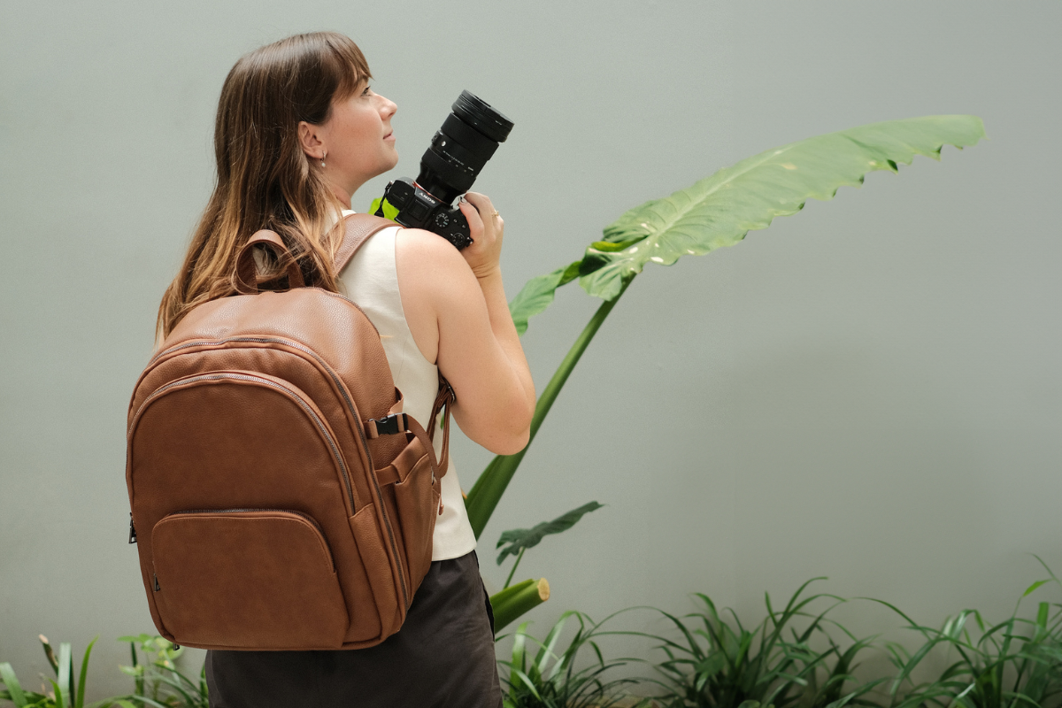 Woman wearing a tan camera backpack holding a camera, standing among plants.