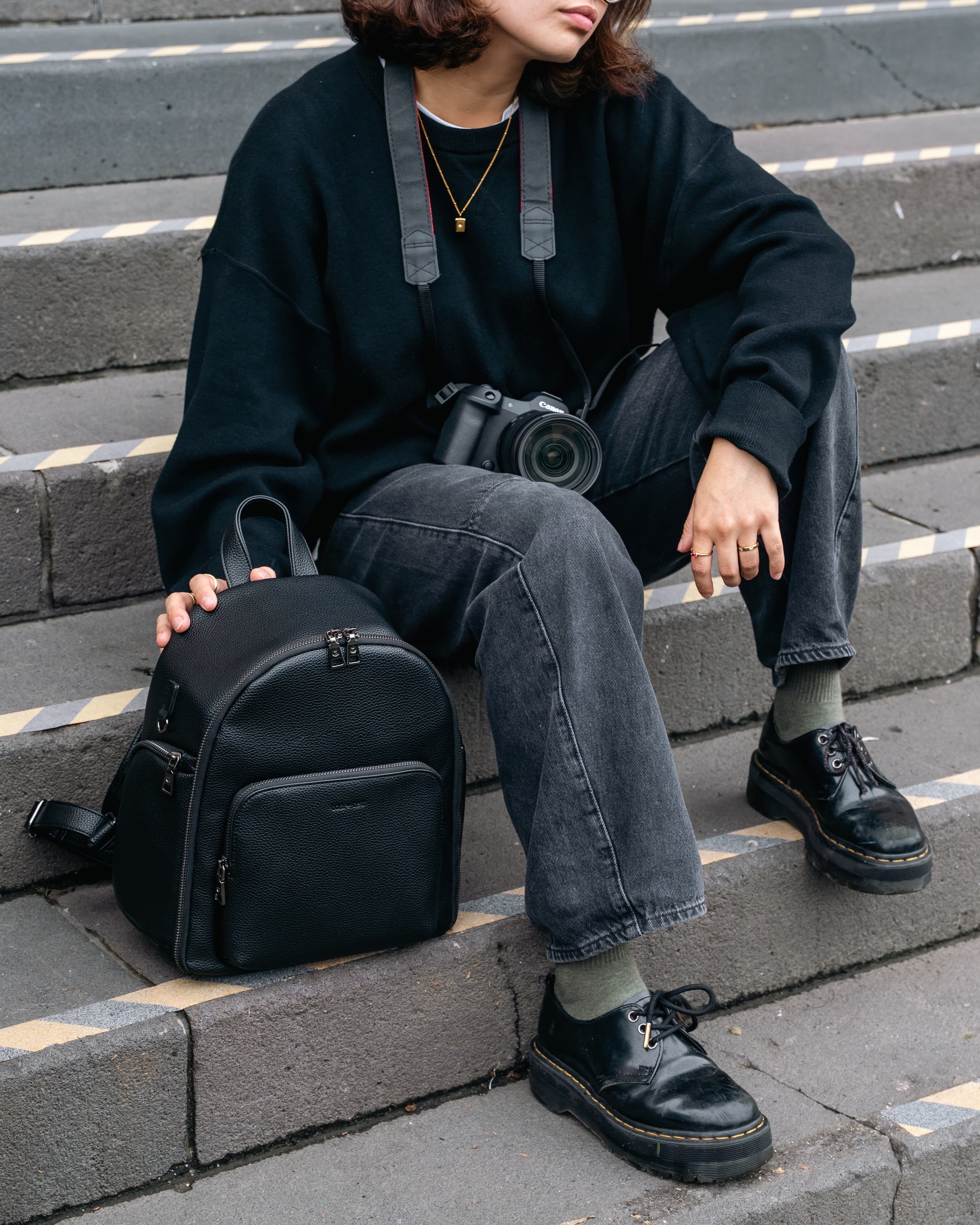 A person sitting on outdoor concrete steps wears a black oversized sweatshirt, dark gray jeans, black shoes, and holds a black backpack next to them. A camera hangs around the neck.
